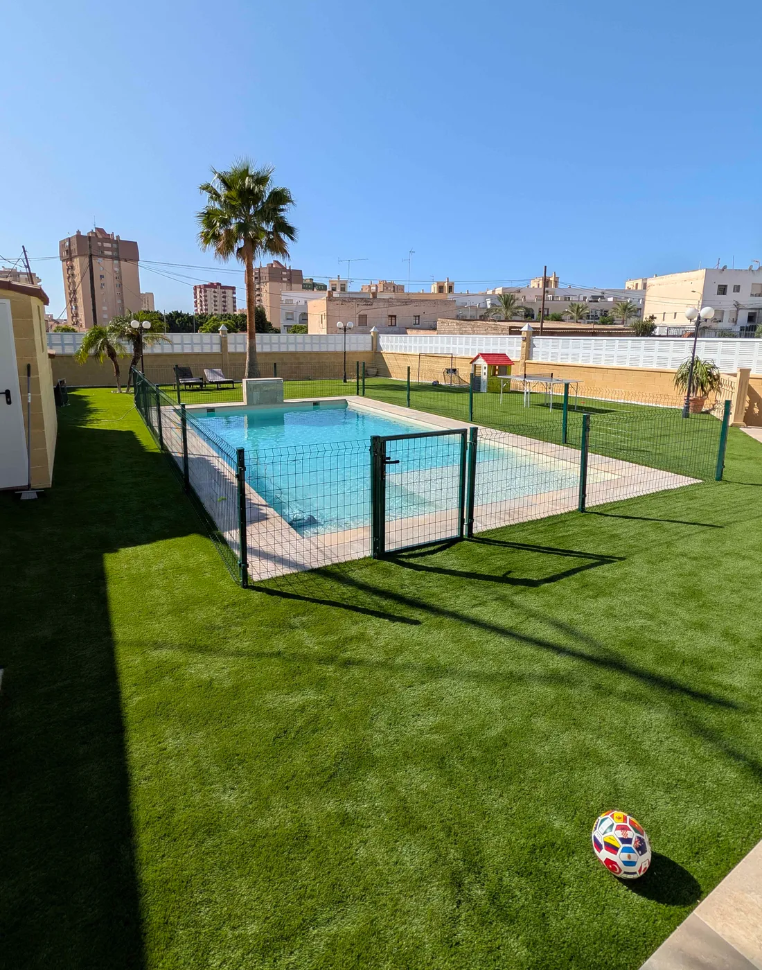 Zona de piscina con vallado rodeada de césped artificial y una palmera